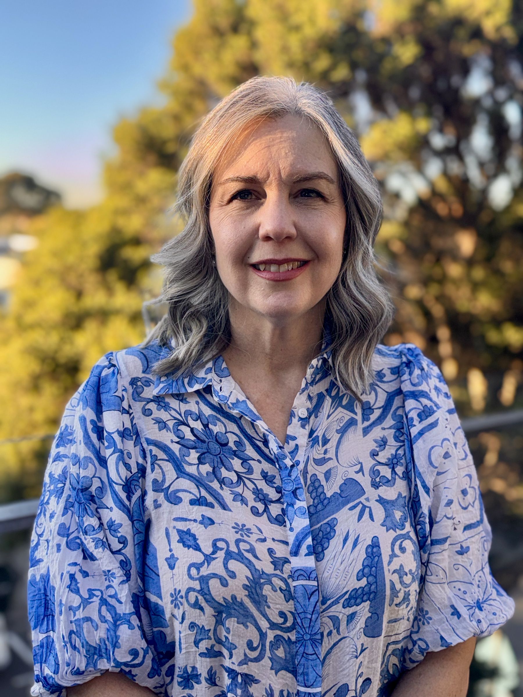 Sharon Peeters, smiling, in a blue-and-white patterned blouse, photographed outdoors among trees in late-afternoon light.
